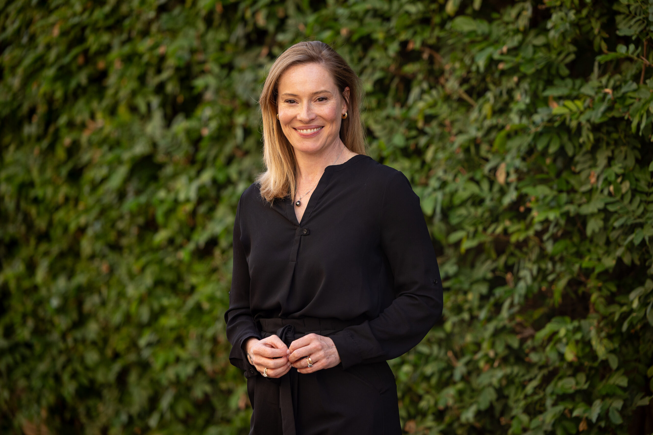 Kelli Fuery, Ph.D., smiles while standing outdoors in front of a lush green ivy-covered wall, wearing a black long-sleeve blouse.